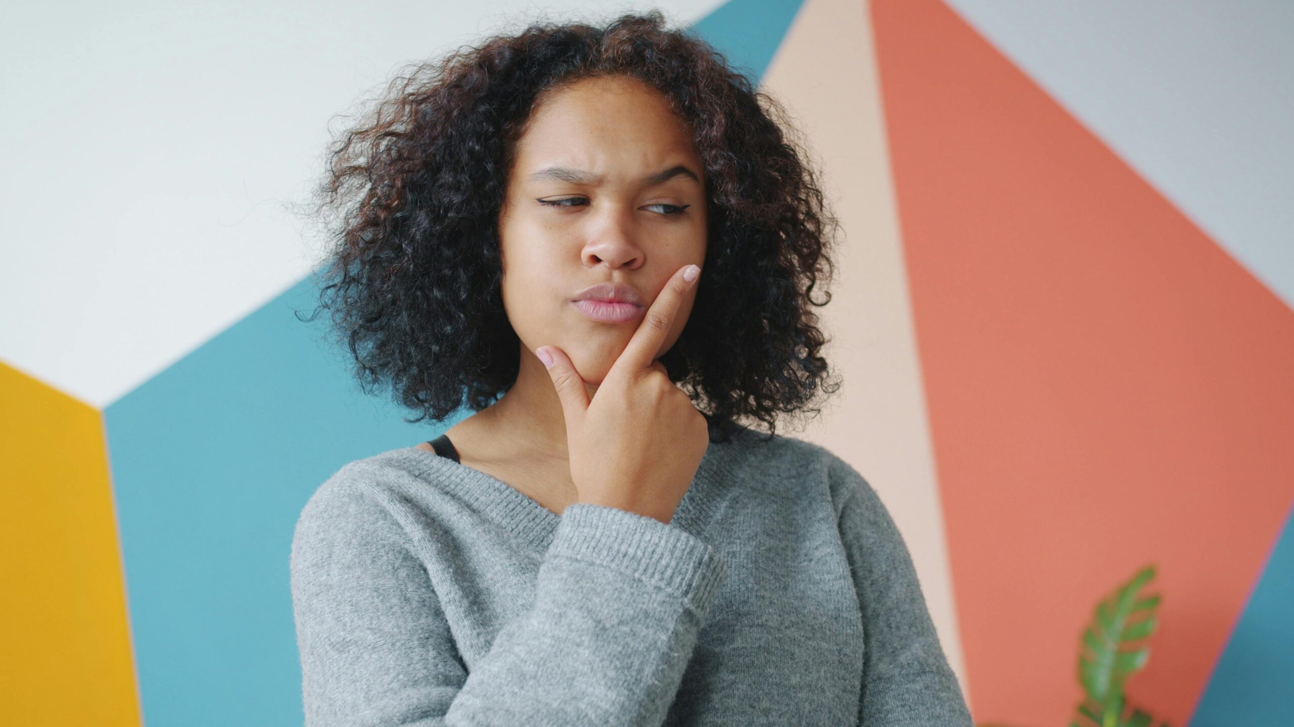 Pensive woman with curly hair pondering in front of colorful geometric wall.
